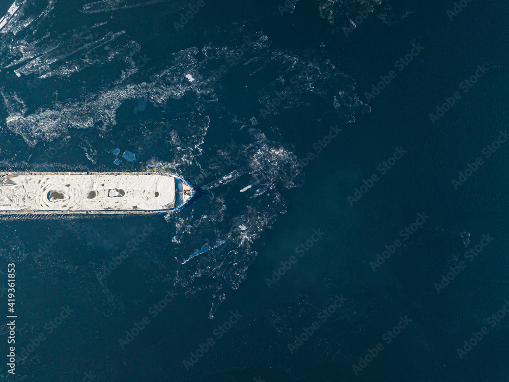 Obraz premium The barge floats on the freezing river. Aerial drone view. Sunny winter day, thin ice on the river.