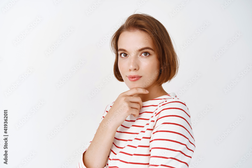 Portrait of thoughtful young woman looking intrigued at camera, touching  face with natural, light make-up, standing on white background Stock Photo  | Adobe Stock