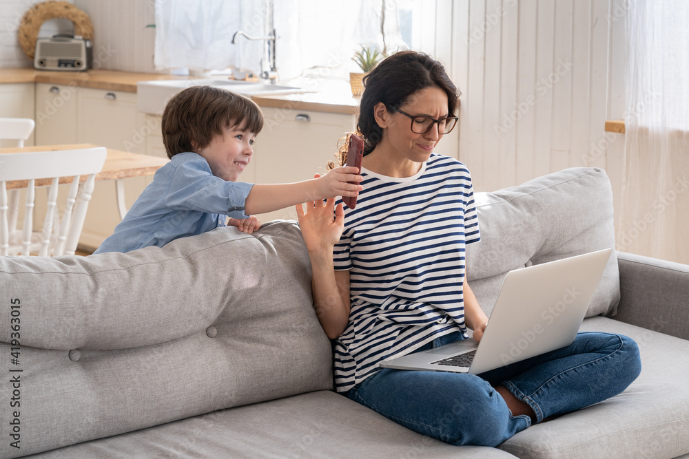 Exhausted freelancer mother sitting on couch at home office during lockdown, working on laptop. Little child distracts from work, shows her smartphone, making noise and asking attention from busy mom. Exhausted freelancer mother sitting on couch at home office during lockdown, working on laptop. Little child distracts from work, shows her smartphone, making noise and asking attention from busy mom.