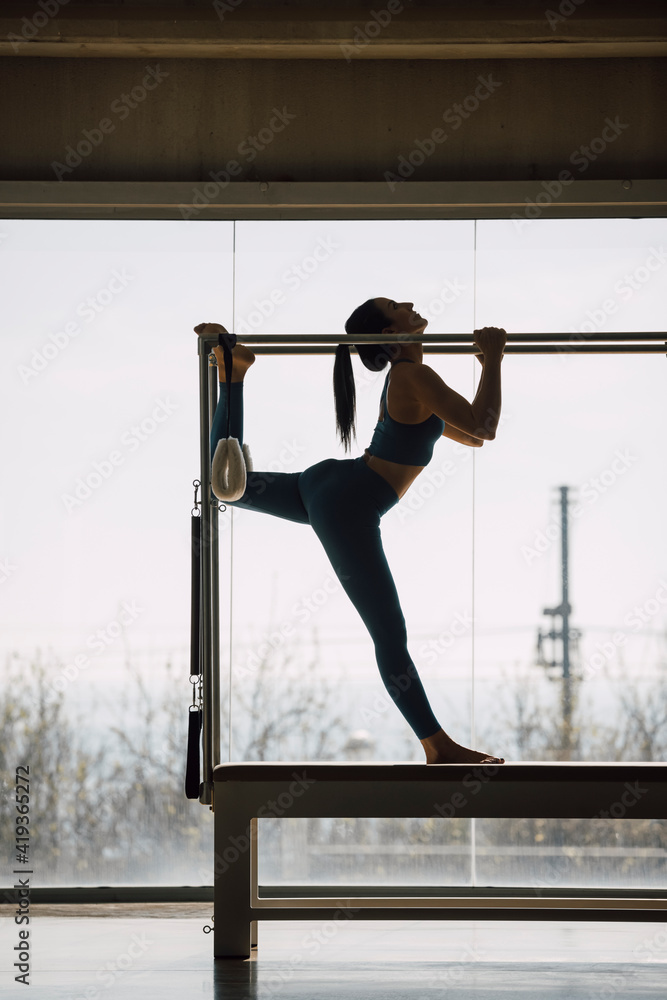 Full length shot of a female Pilates instructor realizing stretching routine, on a Cadillac Machine, Trapeze Table in a big Fitness Studio with wide windows and outside views. Backlight, silhouette.