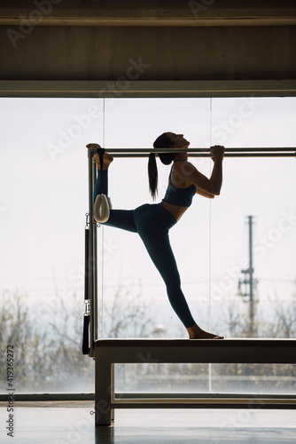 Full length shot of a female Pilates instructor realizing stretching routine, on a Cadillac Machine, Trapeze Table in a big Fitness Studio with wide windows and outside views. Backlight, silhouette.