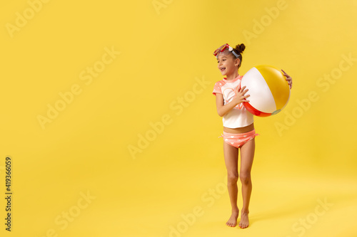 super excited little child girl in swimsuit with beach ball jumping on yellow background. copy space