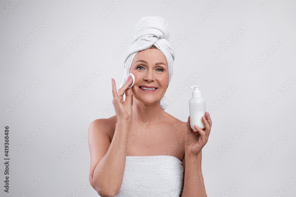 Attractive senior lady applying moisturizing face tonic, looking at camera and smiling on light studio background Attractive senior lady applying moisturizing face tonic, looking at camera and smiling on light studio background