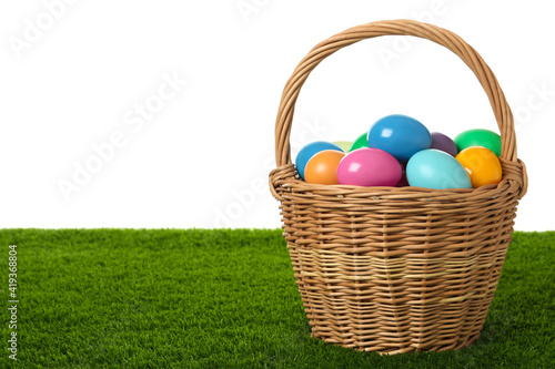 Wicker basket with bright painted Easter eggs on green grass against white background. Space for text