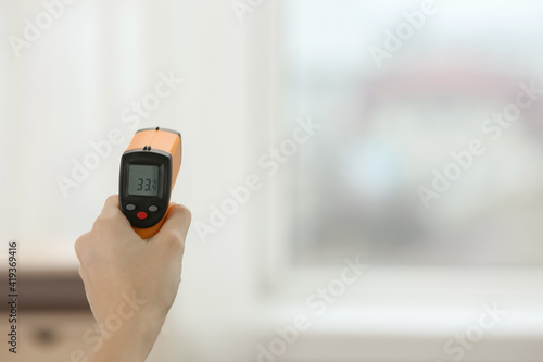 Closeup view of woman with infrared thermometer on blurred background, space for text. Checking temperature during Covid-19 pandemic