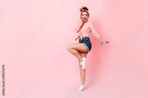 Lady in pink shirt, denim shorts and sneakers jumps up and looks at camera with surprise