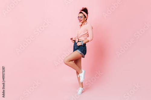 Stylish pin-up girl with red lips lifts her leg on pink background. Full-length shot of woman in pink shirt and denim shorts