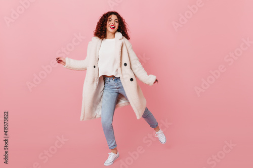 Girl with red lips is dancing on pink background. Woman with dark curly hair dressed in warm fur coat and denim skinny pants smiling