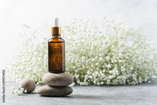 Glass cosmetic bottle with dropper on pebble podium with flowers in the background. Natural background for product presentation