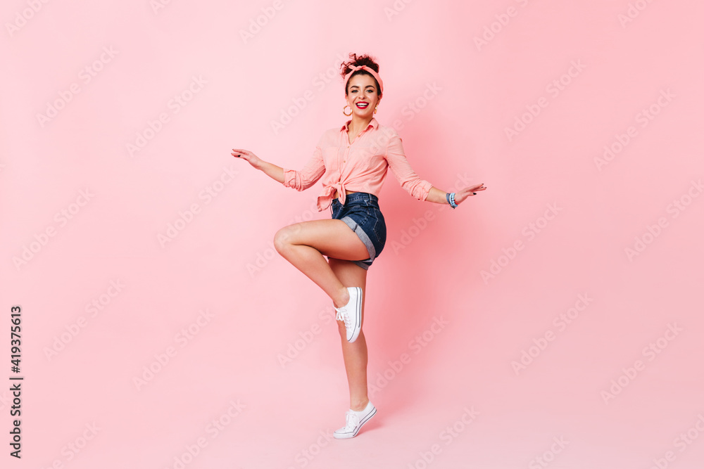 Full-length shot of active woman with bun in pink bandage on her head and dressed in pin-up style clothes dancing on pink background Full-length shot of active woman with bun in pink bandage on her head and dressed in pin-up style clothes dancing on pink background