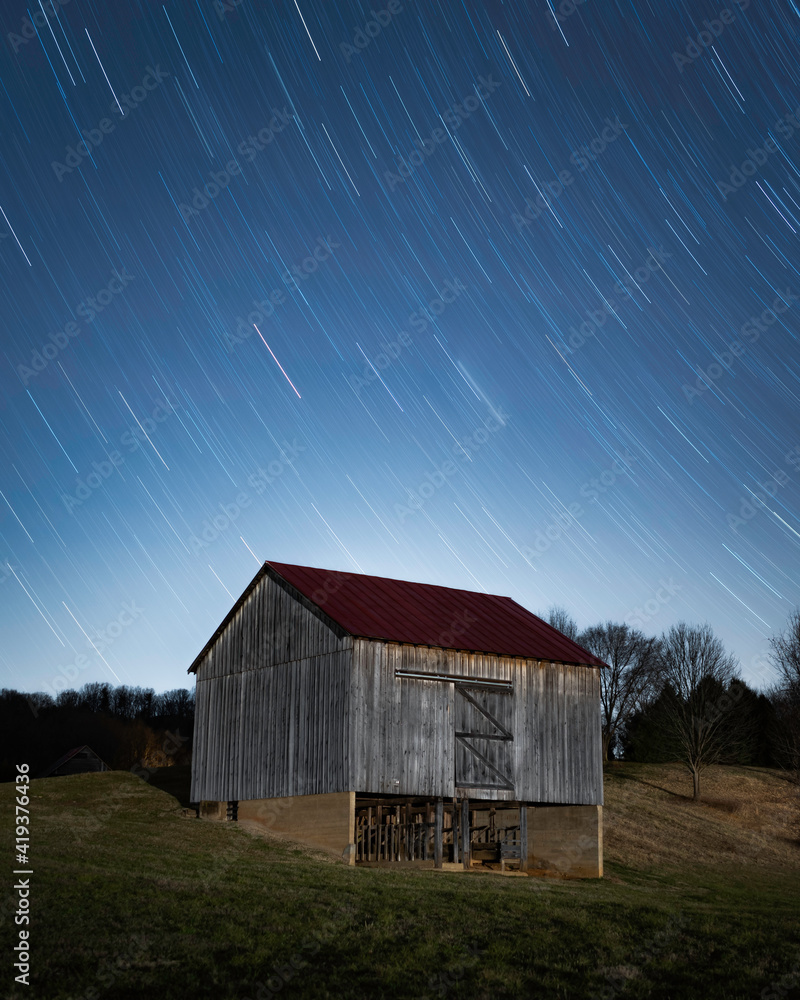 Obraz premium An old barn sits below a night sky as stars zip by in a long exposure.