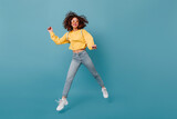Cheerful lady with sincere smile jumping on blue background. Girl with curly hair dressed in cropped sweatshirt and jeans laughs
