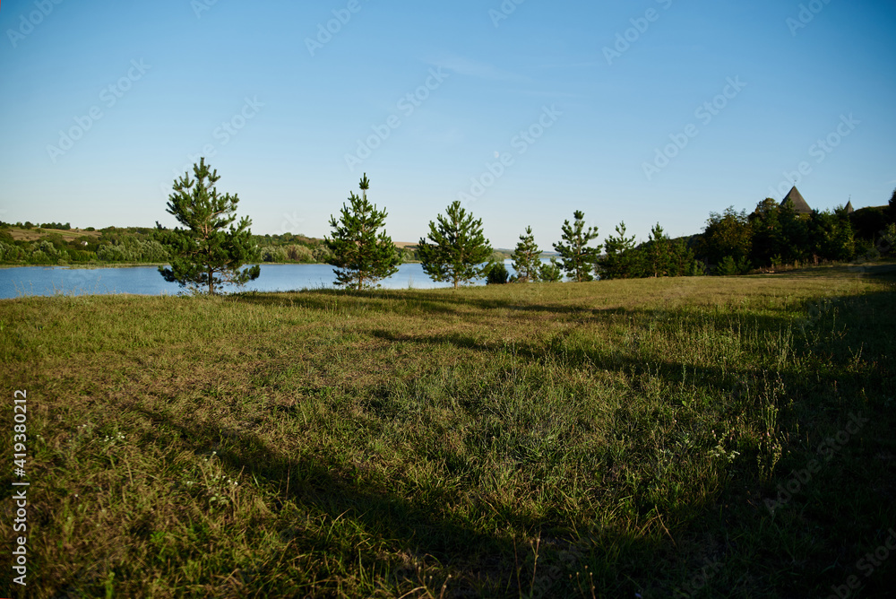 Obraz premium Scenic view of green spruce garden and blue river. Beautiful natural countryside landscape with blue sunny sky. Summer getaway vacation into isolated places. Environmental protection concept.
