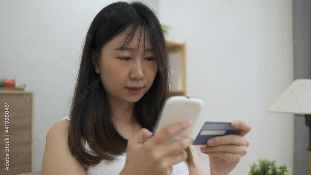 happy asian woman shopping online is smiling while paying by keying in the credit card information on her mobile phone in the living room at home. happy asian woman shopping online is smiling while paying by keying in the credit card information on her mobile phone in the living room at home.
