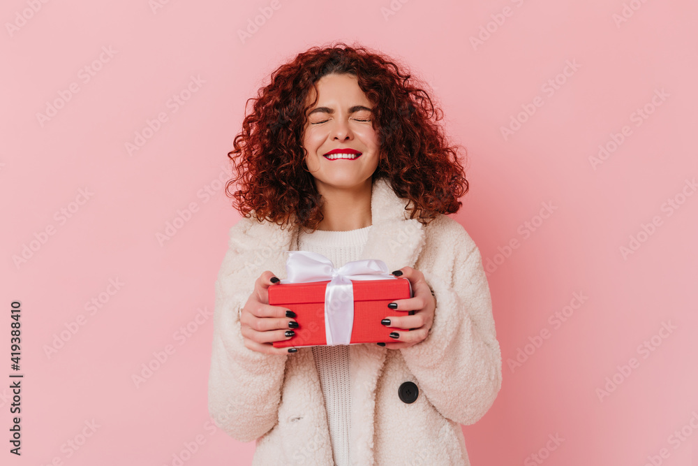 Fototapeta premium Woman in joyful anticipation holds gift. Photo of lady with dark curly hair on pink background