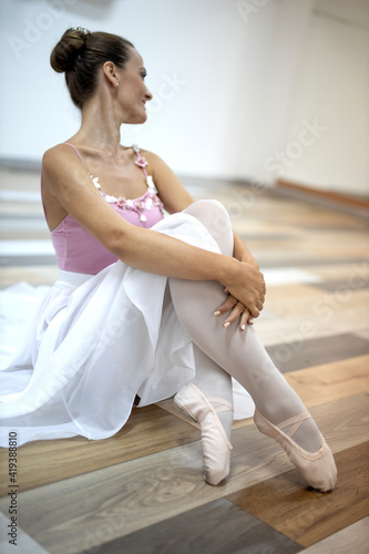 Beautiful professional ballerina in pink dress smiling and sitting on wooden floor in ballet studio