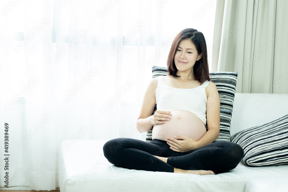 A beautiful Asian woman sitting on a sofa and touching her belly. Pregnancy and expectation concept.