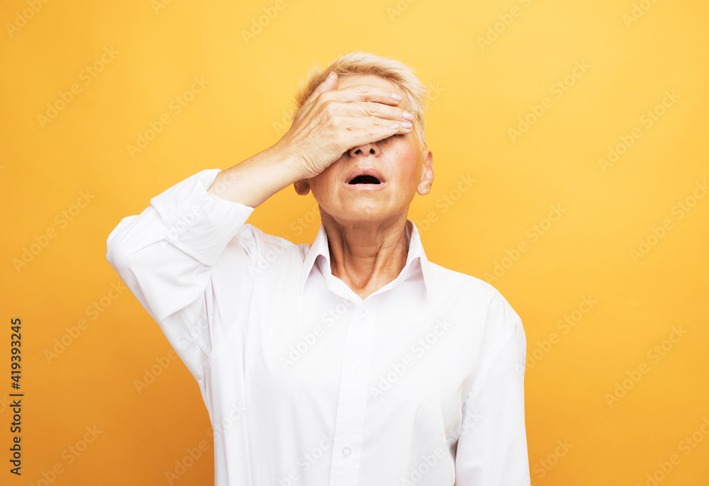 Elderly grey-haired woman in white blouse covering her eyes with hand standing against yellow background Elderly grey-haired woman in white blouse covering her eyes with hand standing against yellow background