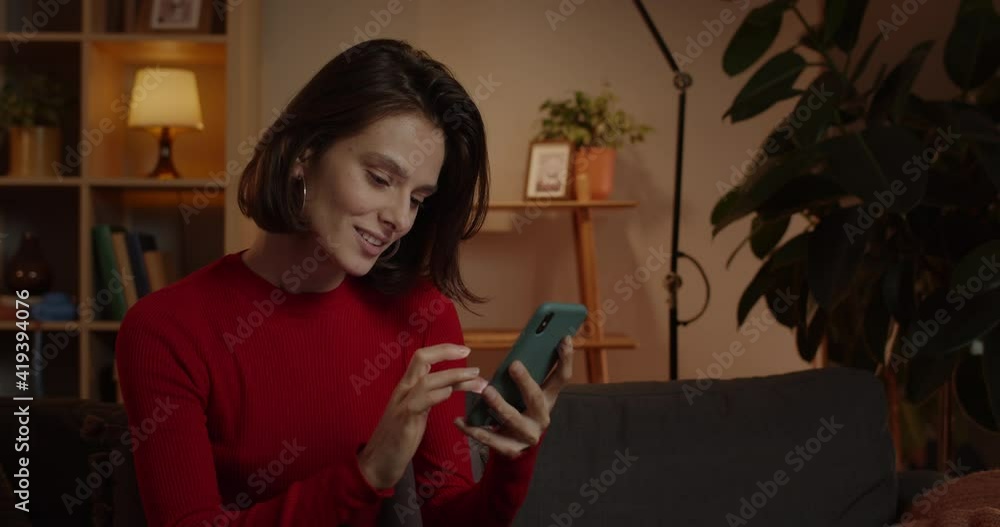 Crop view of seductive woman using smartphone and smiling while sitting on couch at home. Brunette millennial female person scrolling news feed and looking at phone screen Crop view of seductive woman using smartphone and smiling while sitting on couch at home. Brunette millennial female person scrolling news feed and looking at phone screen