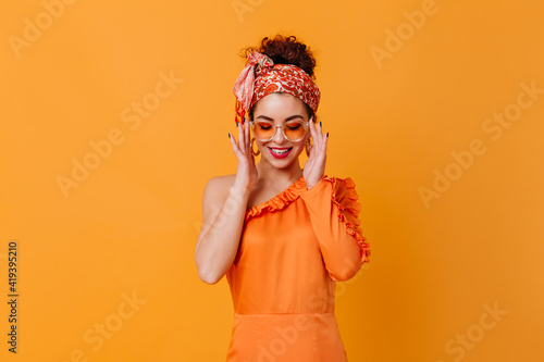 Cute woman in orange glasses, silk dress and headband is smiling on orange background
