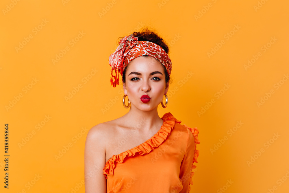 Beautiful blue-eyed woman in orange silk outfit and stylish headband blows air kiss on isolated background Beautiful blue-eyed woman in orange silk outfit and stylish headband blows air kiss on isolated background