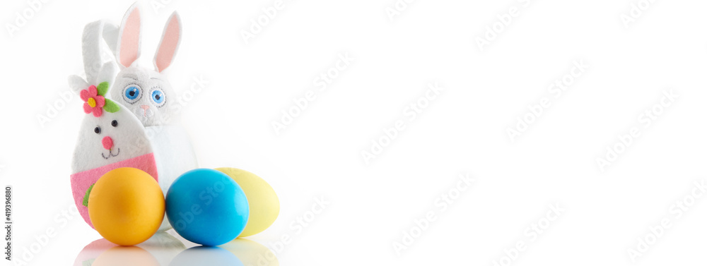 Fototapeta premium Banner with hand made white rabbit in basket and three colored eggs on white background.