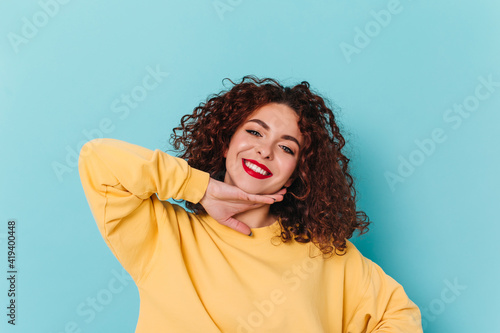 Close-up portrait of blue-eyed girl with red lips. Curly woman in yellow sweater touches her chin and poses on blue background © Look!