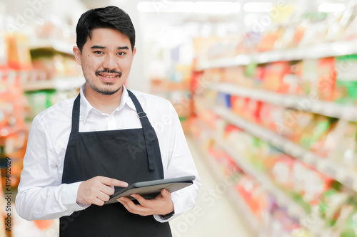 close up adult Middle East business owner man holding tablet to check about inventory stock of goods or online order in supermarket isolated on blur market background for SME concept