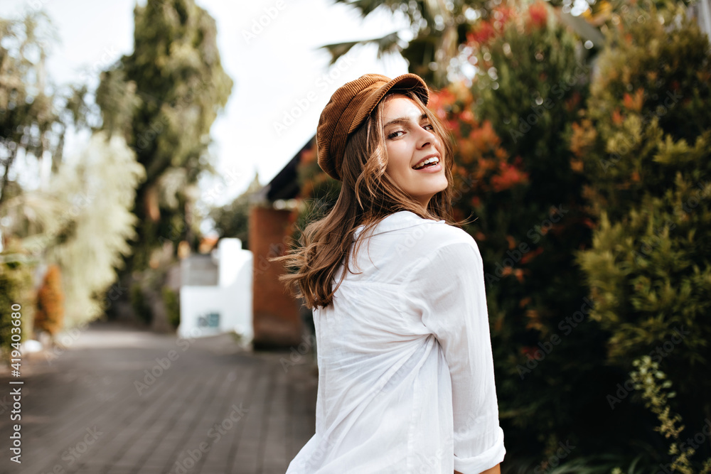 Dark blonde cheerful girl in brown cap and white cotton blouse walks along path in park