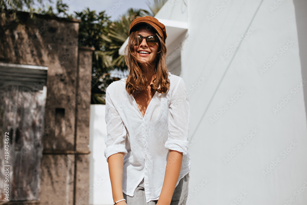 Cheerful lady with short hair and tattoo on her arm is smiling against background of one-story buildings. Shot of lady in white blouse, gray shorts and glasses Cheerful lady with short hair and tattoo on her arm is smiling against background of one-story buildings. Shot of lady in white blouse, gray shorts and glasses