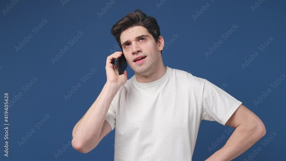 Dissatisfied irritated angry young man 20s in white t-shirt posing isolated on blue background in studio. People lifestyle concept. Talking on mobile cell phone spreading hands screaming swearing Dissatisfied irritated angry young man 20s in white t-shirt posing isolated on blue background in studio. People lifestyle concept. Talking on mobile cell phone spreading hands screaming swearing