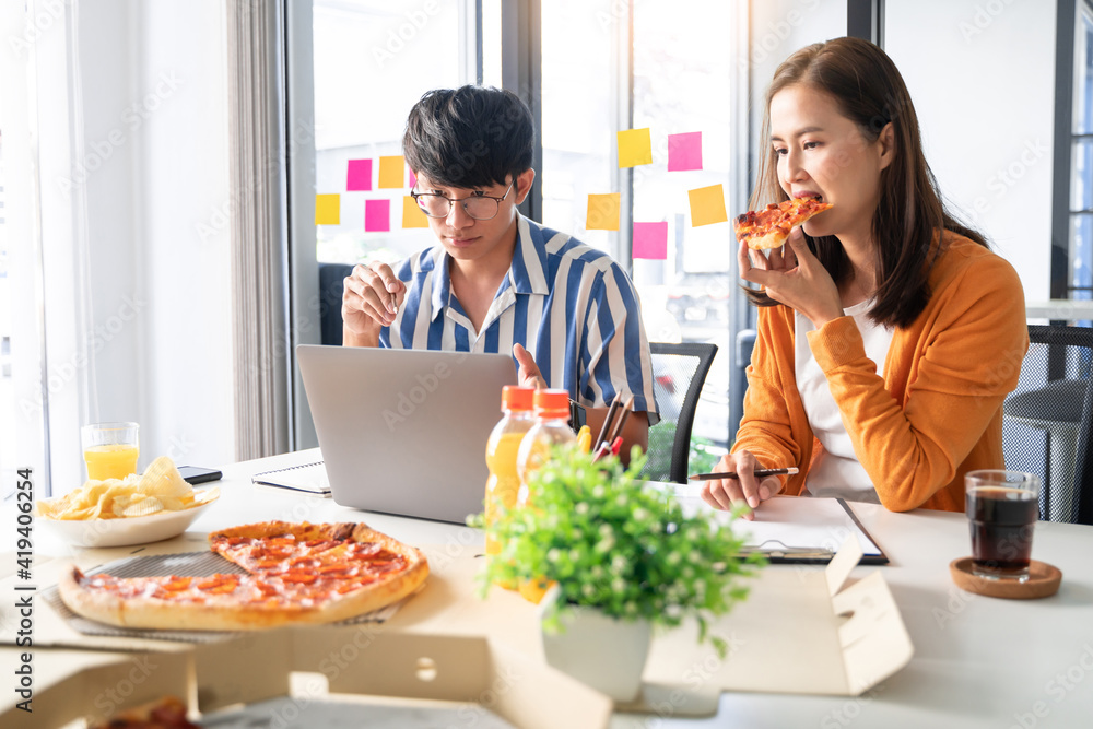 Friendly asian business team colleagues eating pizza and potato chips together in office have fun talk enjoy corporate party