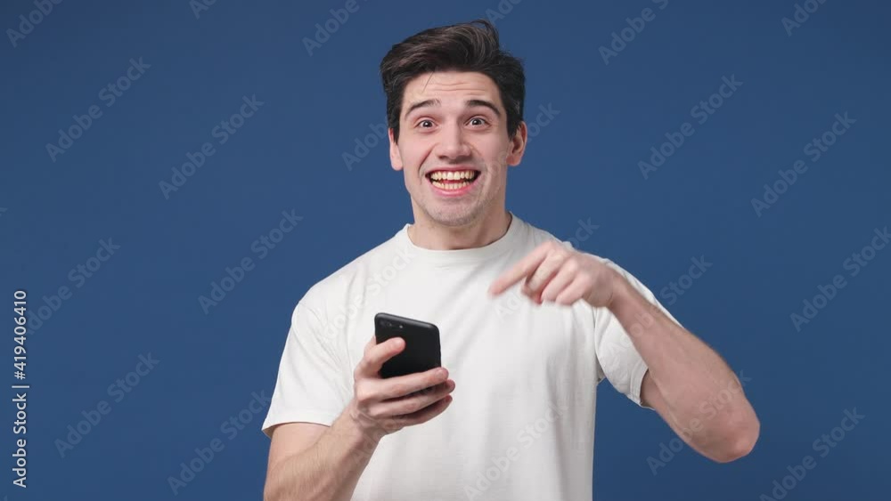 Shocked surprised young man 20s in white t-shirt posing isolated on blue background studio. People lifestyle concept. Pointing index finger on mobile cell phone screaming doing winner gesture thumb up Shocked surprised young man 20s in white t-shirt posing isolated on blue background studio. People lifestyle concept. Pointing index finger on mobile cell phone screaming doing winner gesture thumb up