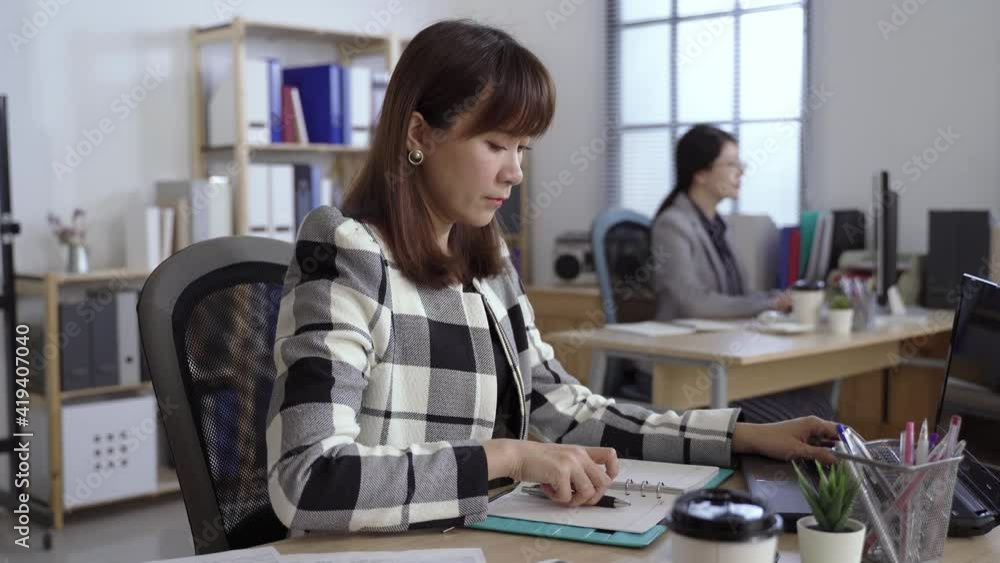 pretty asian office lady is taking notes in her notepad while typing reports on the laptop at a bright workplace. pretty asian office lady is taking notes in her notepad while typing reports on the laptop at a bright workplace.