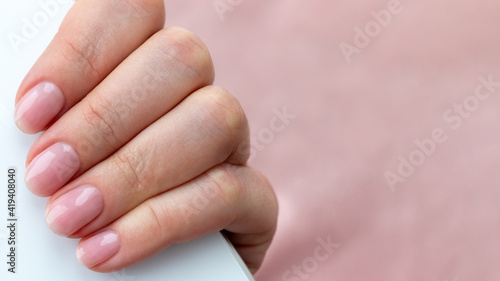 Female hand with beautiful manicure - pink nude nails with white smartphone on pale pink background, closeup with copy space. Selective focus