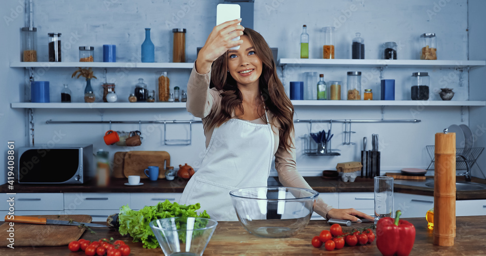 happy woman in apron taking selfie near ingredients on table