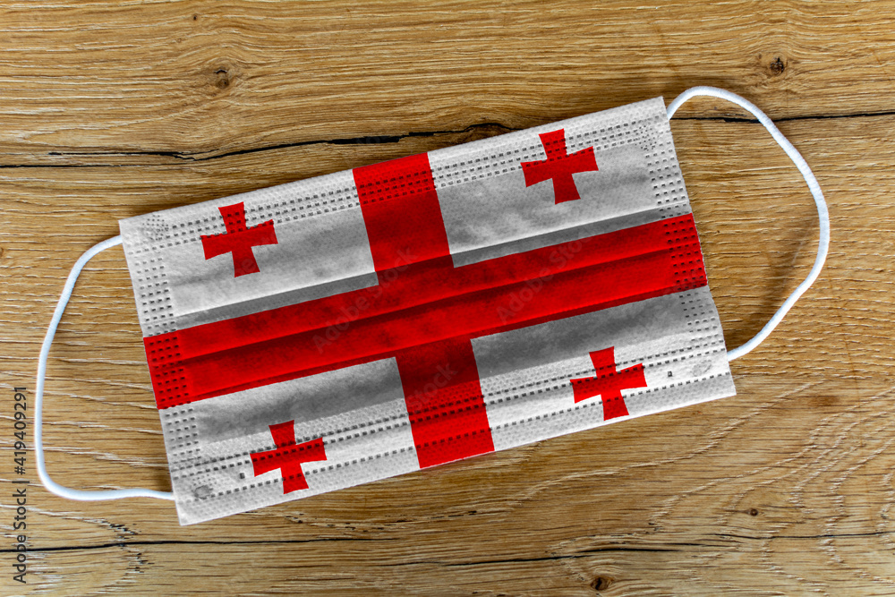 Fototapeta premium A surgical face mask with the painted flag of Georgia on a wooden background. Concept of Georgia during a pandemic, Covid-19, Coronavirus.