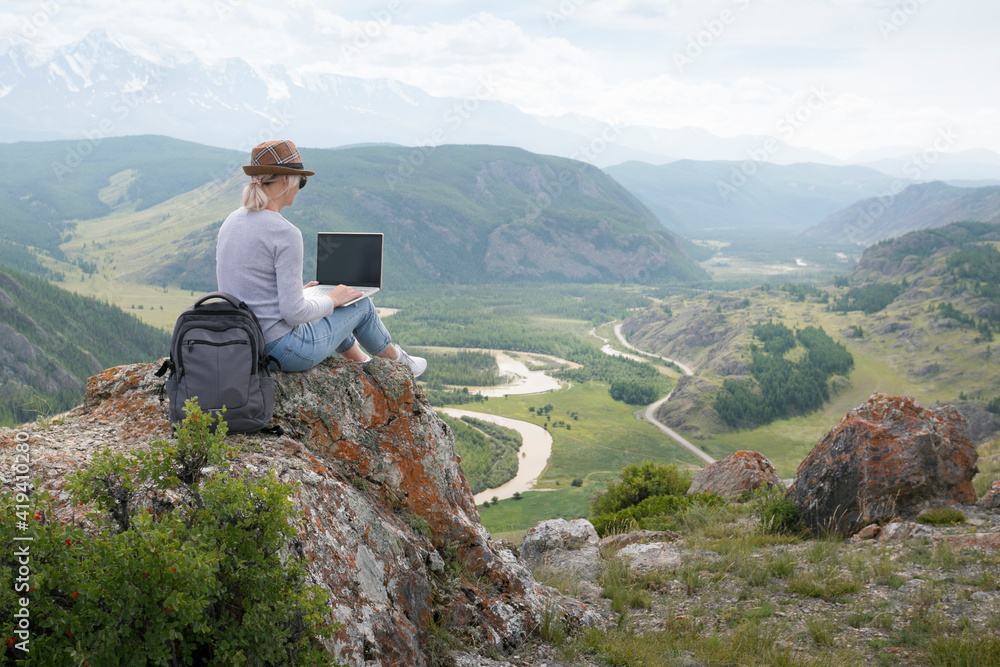 Beautiful woman traveler in a hat in mountains with a laptop. Work outside the office.