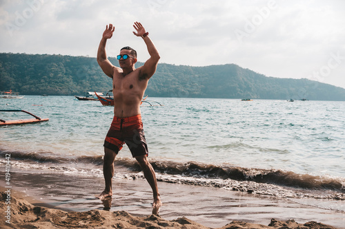 Asian fitness guy doing jumping jacks, star jump or burpees exercise at seaside outdoors. Healthy lifestyle by the beach. photo