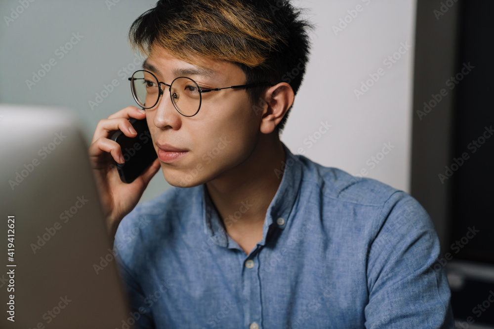 Serious asian guy talking on cellphone while working with laptop Serious asian guy talking on cellphone while working with laptop