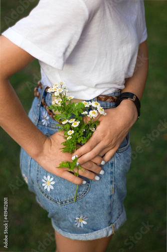 unrecognizable teenager girl holds a small bouquet of daisies near the back pocket of denim shorts. Close-up without a face. Hello summer. Youth, tenderness, romance