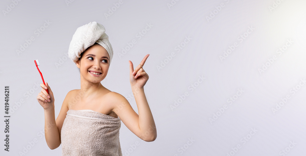 Banner, long format on grey background. Portrait of a smiling pretty woman wearing bath towel, holding toothbrush, looking away, showing forefinger to empty side space for ads Banner, long format on grey background. Portrait of a smiling pretty woman wearing bath towel, holding toothbrush, looking away, showing forefinger to empty side space for ads