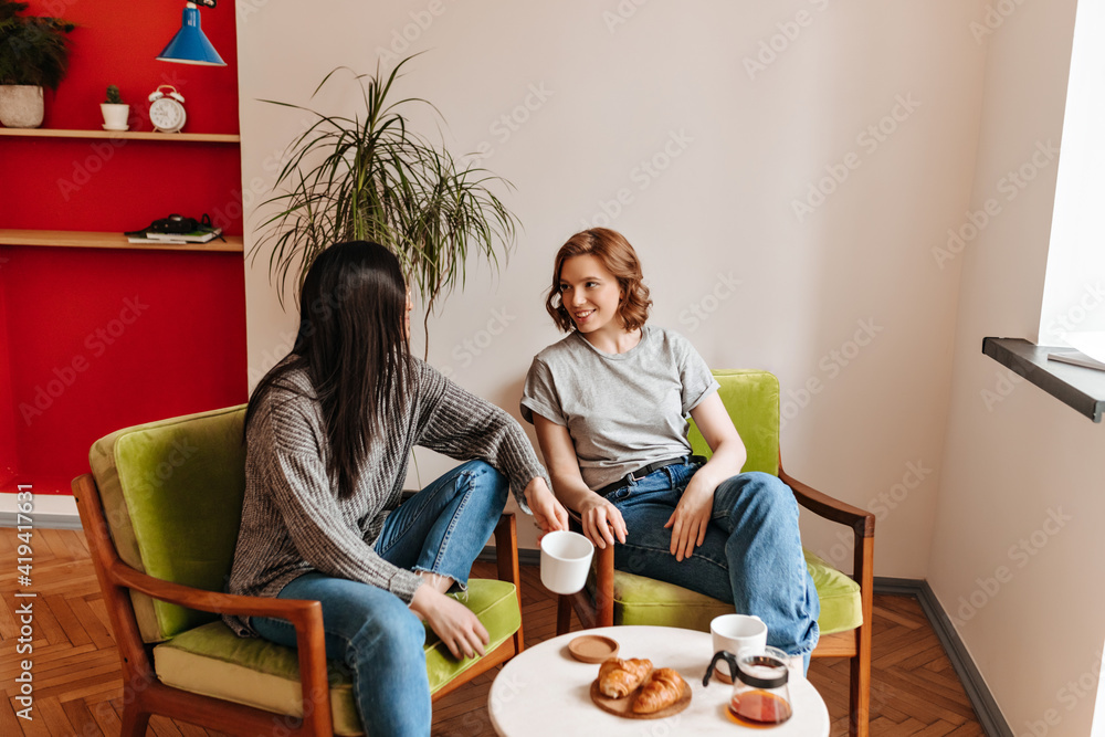 Two girls enjoying tea time at home. Best friends sitting in armchairs and drinking coffee.