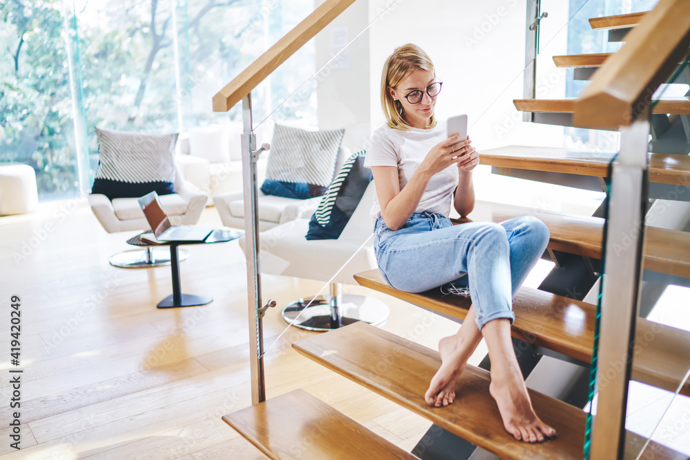 Calm female in casual clothes sitting on stairs looking at screen of smartphone while chatting with friends having free time