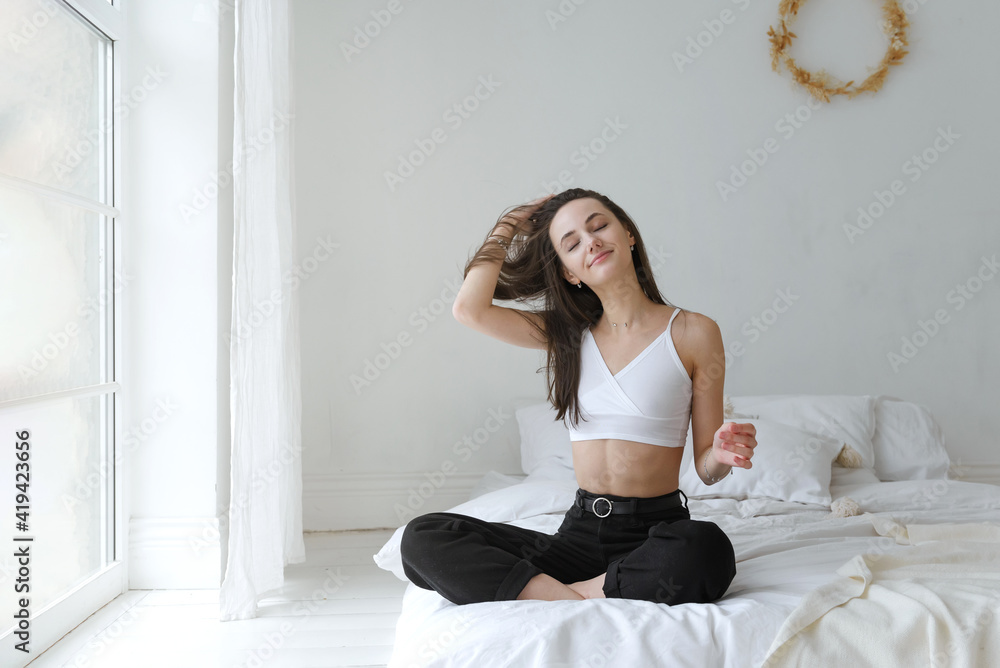 European girl in a white top and black jeans is sitting in a bright room on a bed