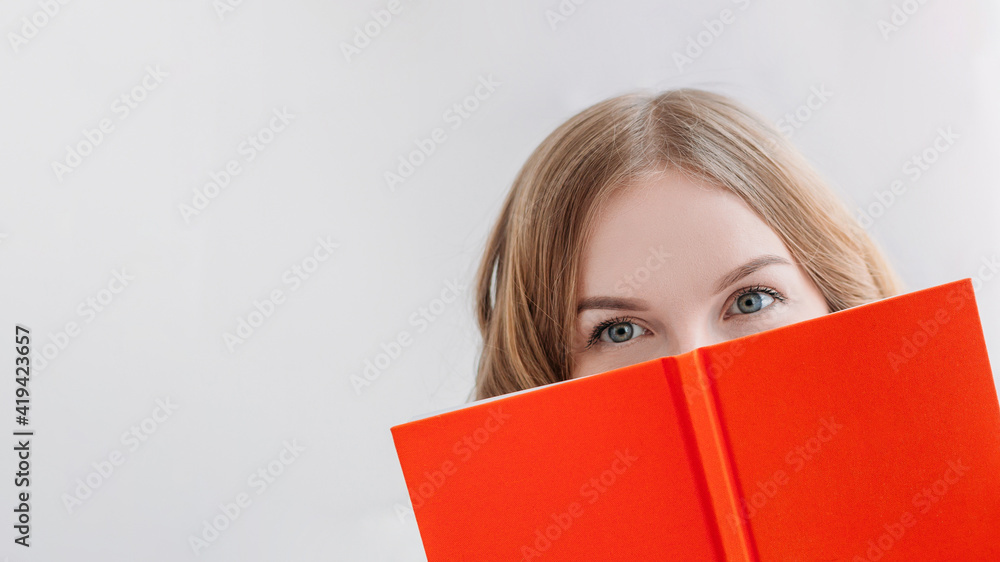 Portrait of a pretty young blond girl hiding behind an open orange book. Happy Woman studying or working at home Portrait of a pretty young blond girl hiding behind an open orange book. Happy Woman studying or working at home