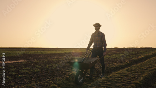 Male farmer pushing wheelbarrow over farmland wearing straw hat plaid shirt rubber boots sunglasses at sunset