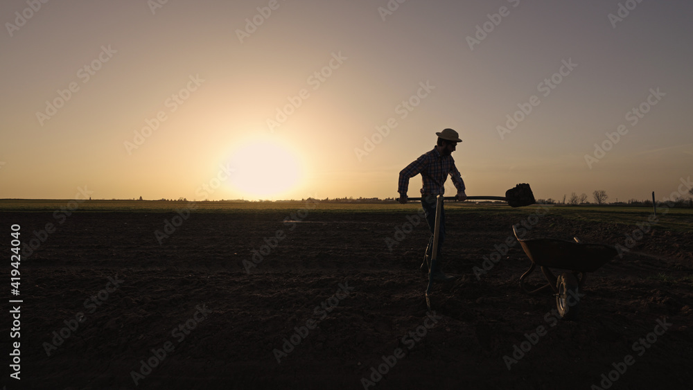 Male farmer shovels earth in wheelbarrow on farmland wearing straw hat plaid shirt rubber boots sunglasses at sunset