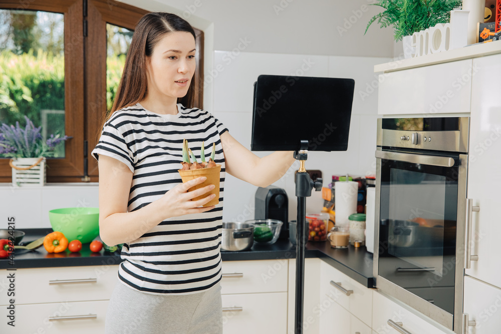 Young woman in kitchen recording video on tablet. Smiling woman working on food blogger concept with vegetables in kitchen. Camera live streaming. woman holding onion in a vase Young woman in kitchen recording video on tablet. Smiling woman working on food blogger concept with vegetables in kitchen. Camera live streaming. woman holding onion in a vase