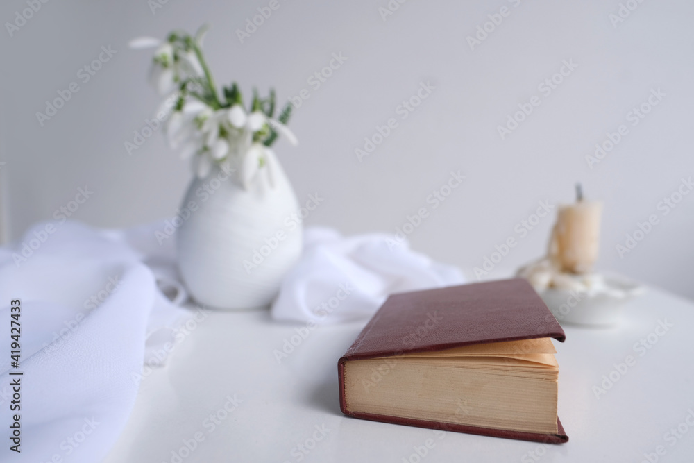 still life in high key style, old paper book in a dark red binding on white table, candle, bouquet of half snowdrops in vase, concept of changing seasons, reading literature
